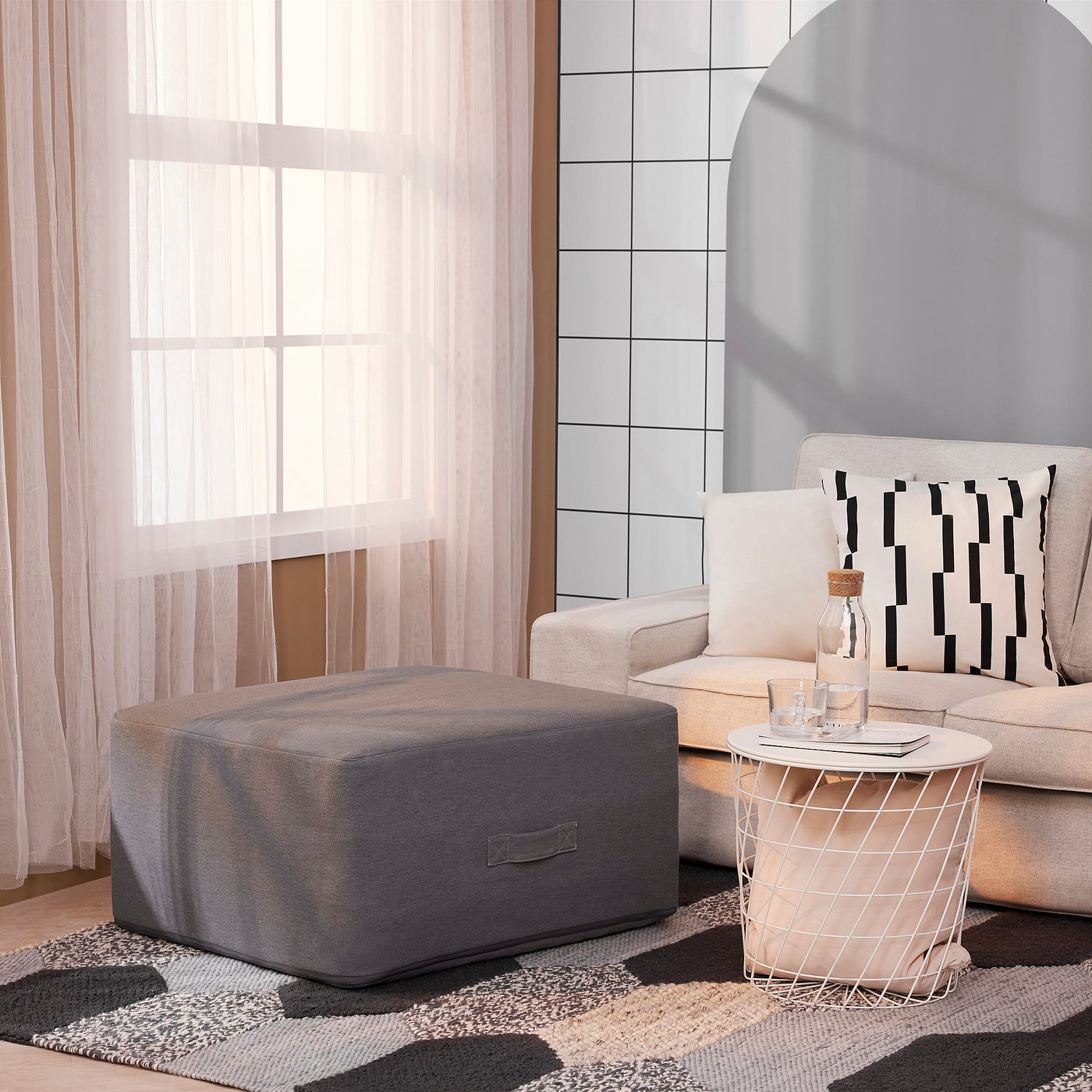 Beige sofa with stripe pillows, grey ottoman, patterned rug, side table with items.