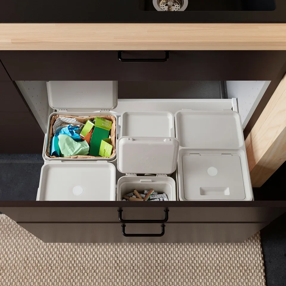 Organised drawer with white HÅLLBAR bins for waste sorting, one containing various items and three empty, mounted under a kitchen counter.