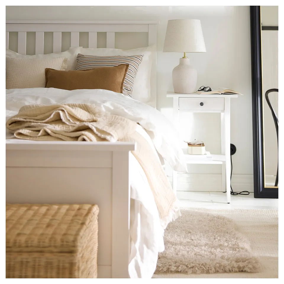 Bedroom with white bed, beige cushions, white rug, woven bench, and small white bedside table with lamp.