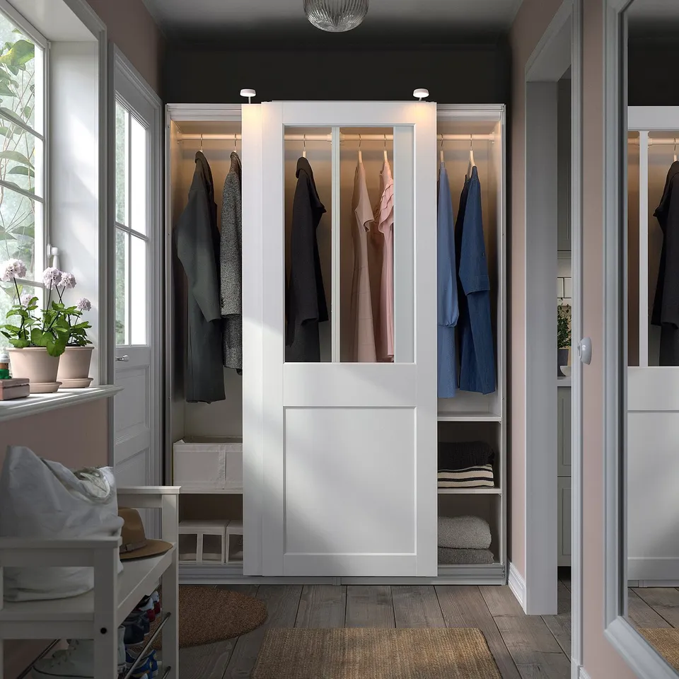 White PAX wardrobe with glass doors showing clothes and towels, beside a window with a plant.
