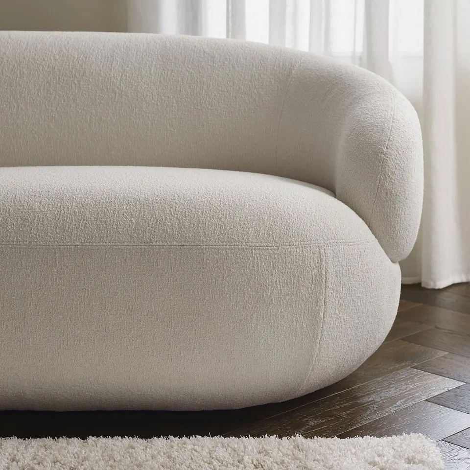 Modern curved sofa with plush, off-white fabric, offering comfort and style. Placed on wooden floor with a cosy rug.