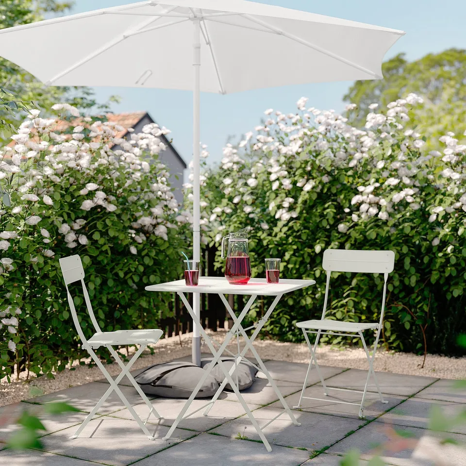 White outdoor table & chairs with red drink set under white umbrella, surrounded by flowers.