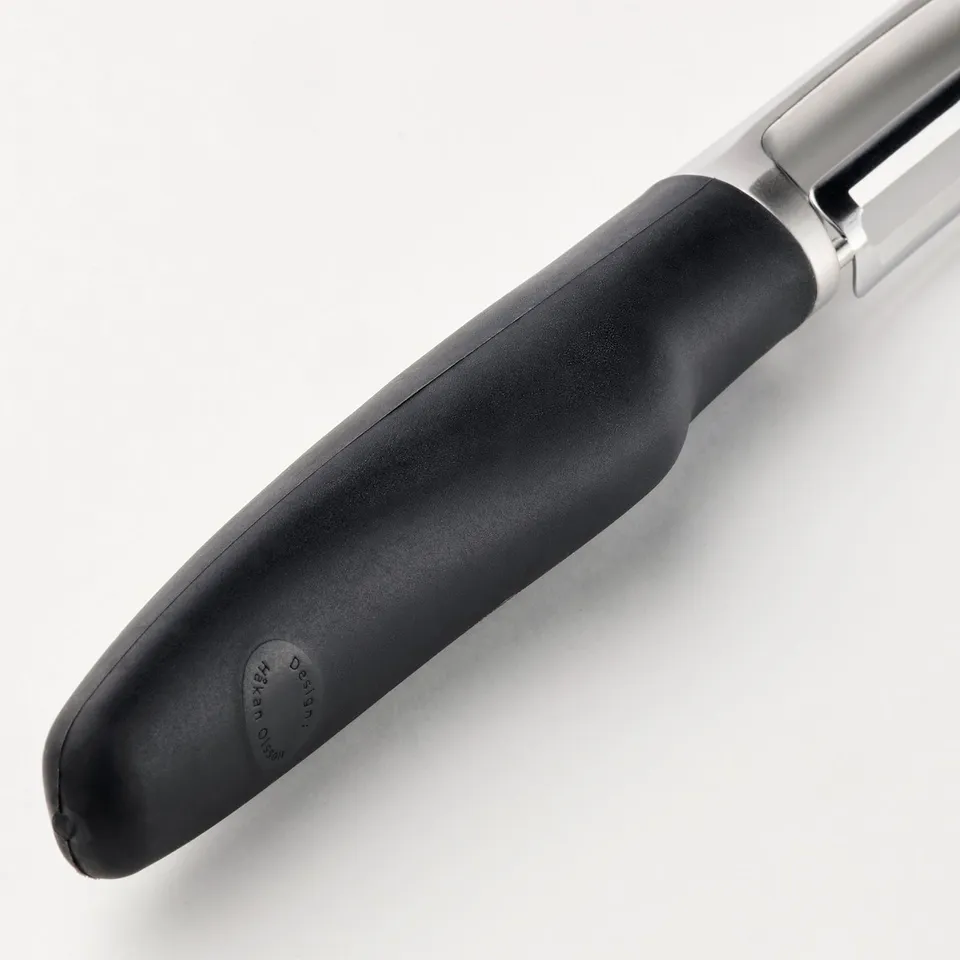 Peeler with black grip and metallic body rests on surface. Firm grip and sharp stainless steel blade.