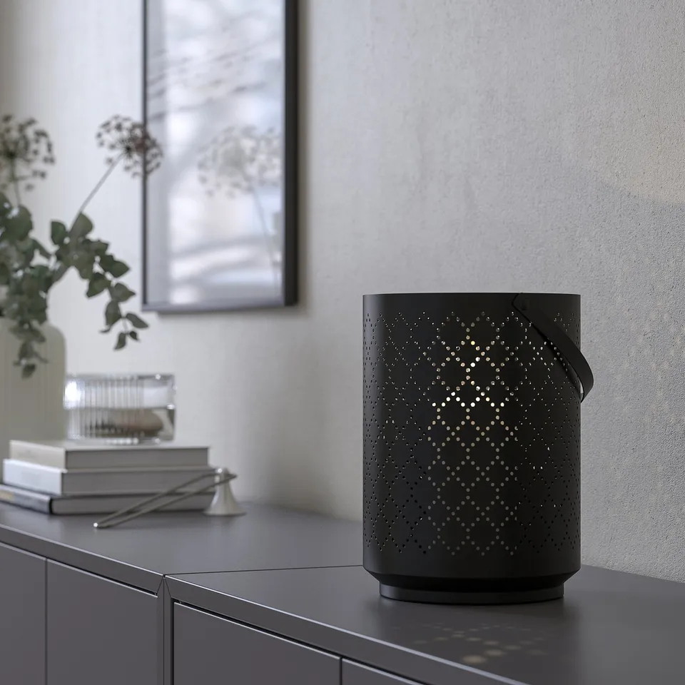 Black patterned lantern on shelf, casting light, with books, scissors, and vase nearby.