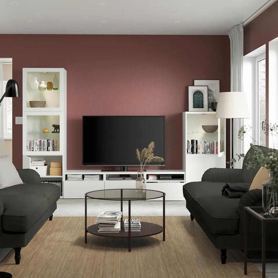 Modern living room with maroon wall, large tv on BESTÅ unit with books, two black sofas facing it, glass coffee table with books and vase.
