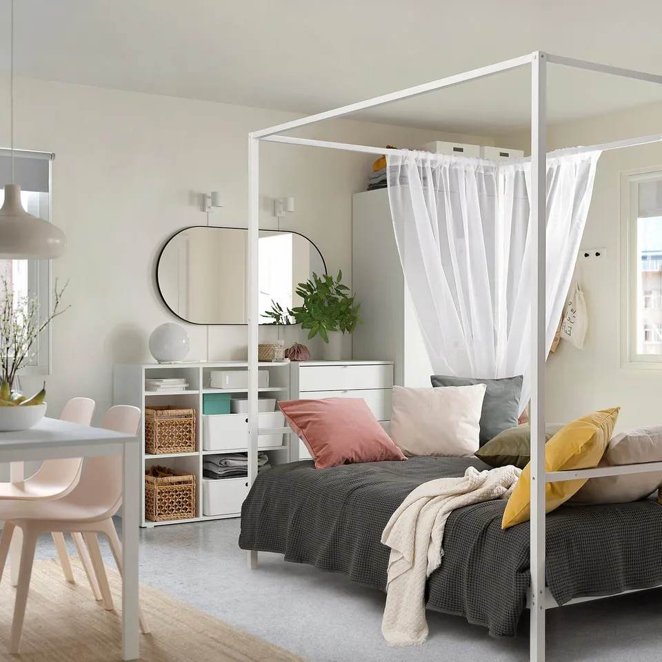 Modern bedroom with VITARNA white metal four-poster bed, grey bedding, colourful pillows, shelving, dining table, and large mirror.