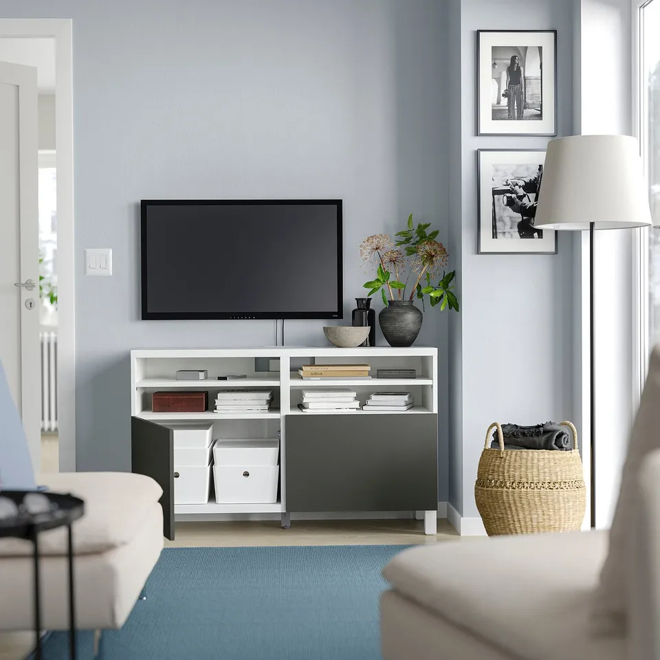 Modern living room with white BESTÅ tv bench, flat screen tv, grey rug, black and white framed art, wicker basket, and white lamp.