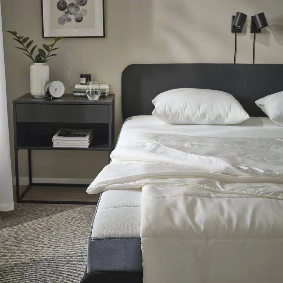 Modern bedroom with black duvet, white linens, bedside table with books, clock & plant.