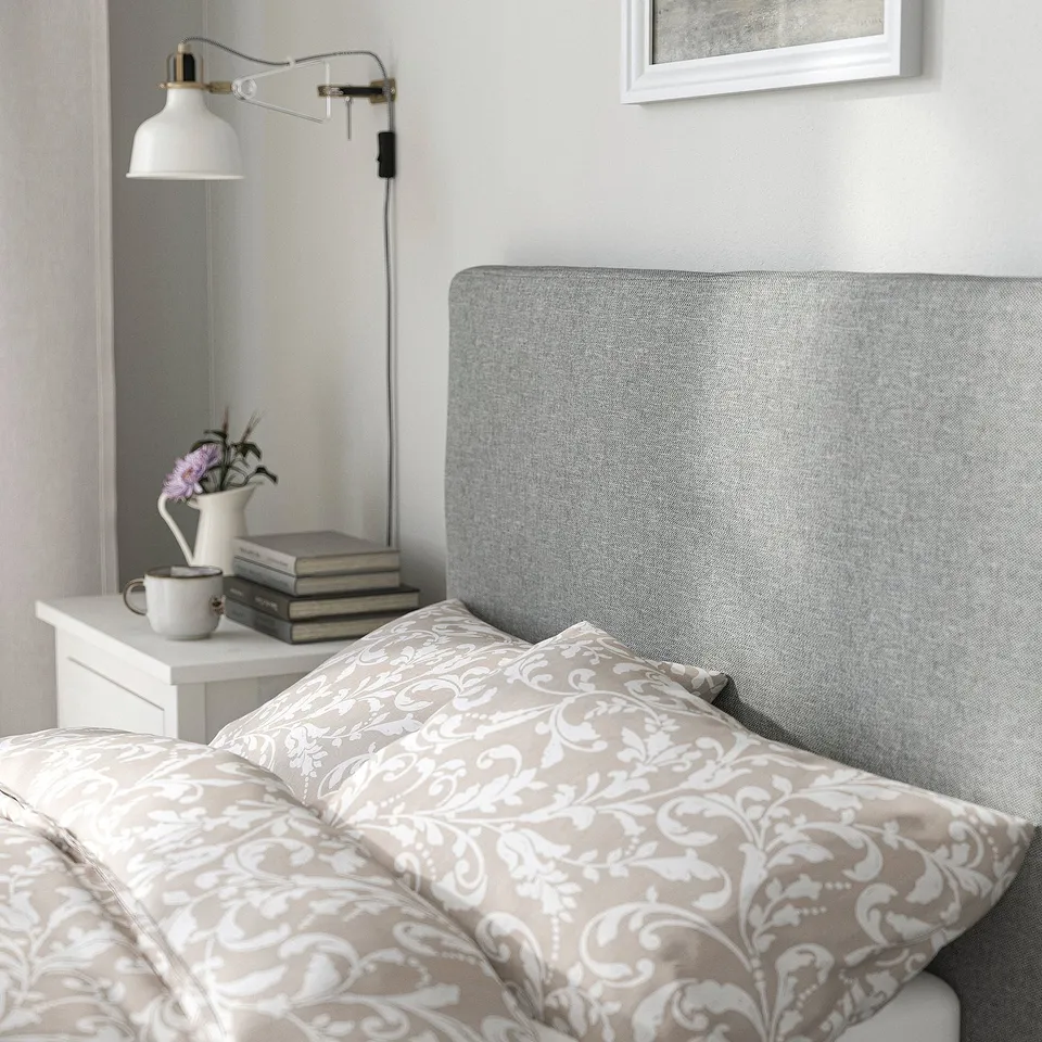 Cosy bed with grey headboard, beige bedding, bedside table with books, flowers, mug, and wall lamp.