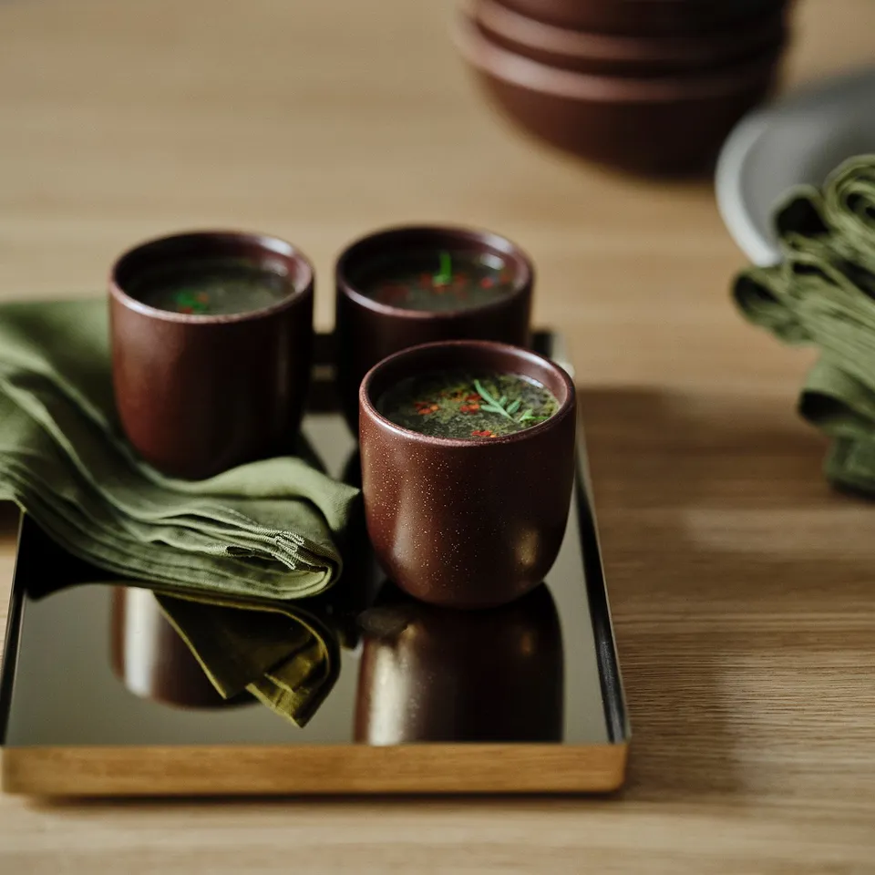Three dark cups on a tray with green linen napkins, filled with a green liquid and garnished with herbs.