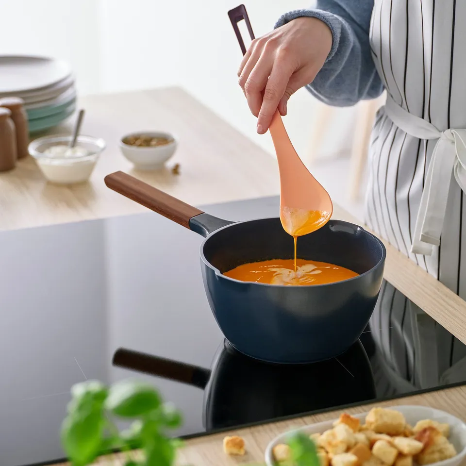 Person stirs orange soup in blue pan using spoon with wooden handle and silicone edge, on kitchen stove.