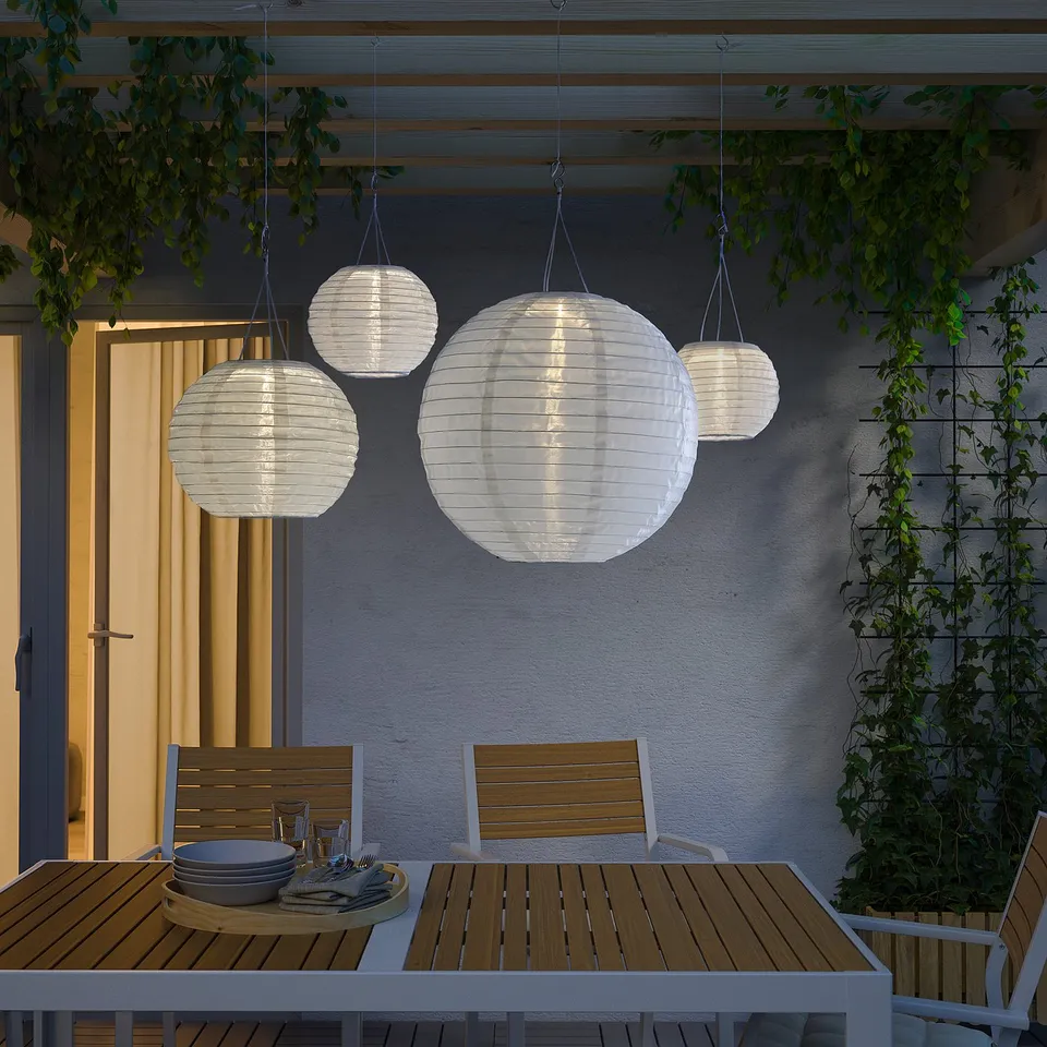 White solar lanterns above wooden table & chairs, pergola with greenery.