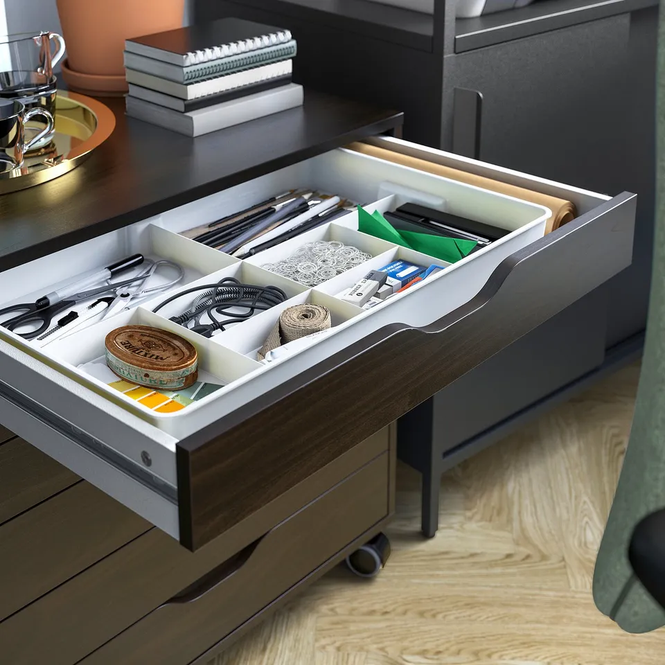 Organised desk drawer with compartments holding various small items like stationery, cables, and tape. Grey ALEX desk, wood floor, green chair.
