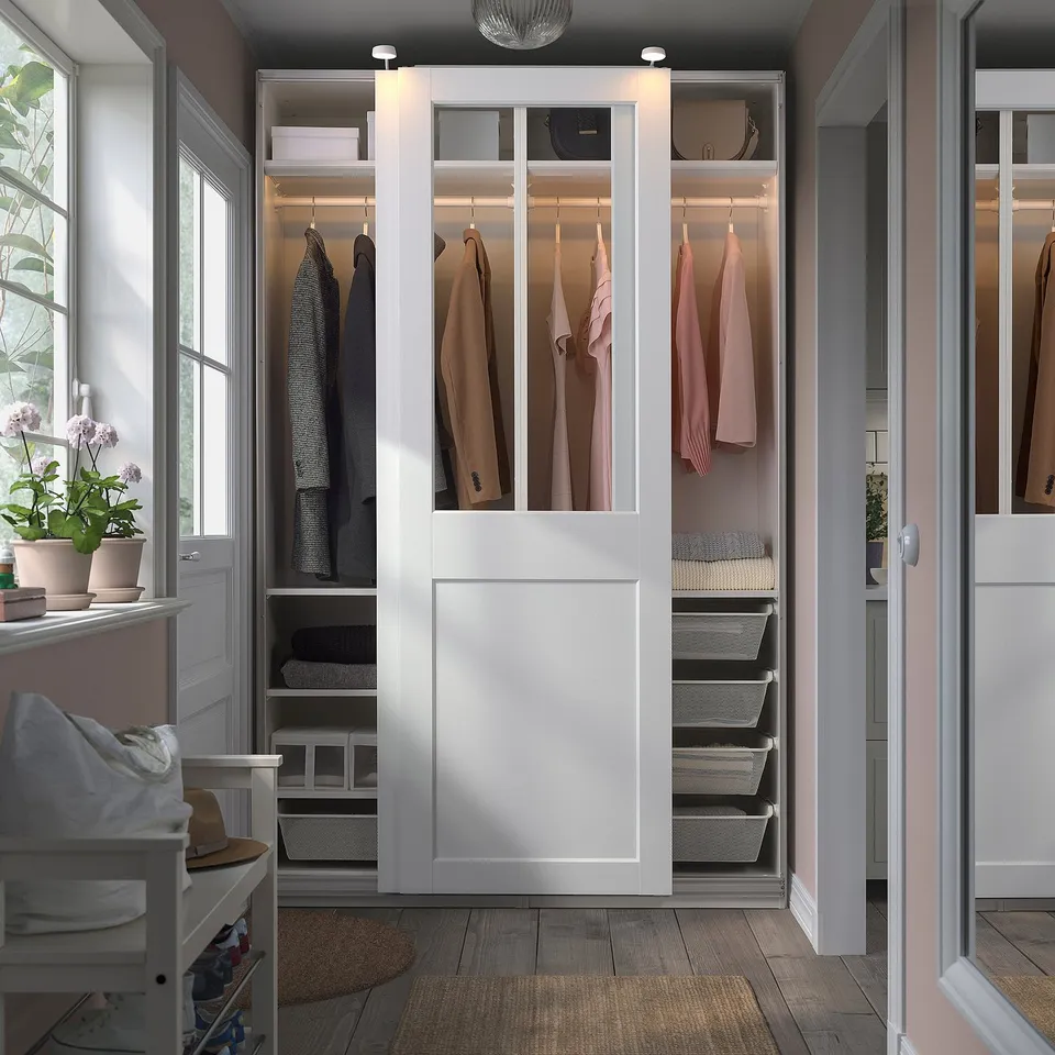A white PAX wardrobe with glass doors, displaying clothes. Its large, surrounding a smaller white chair, and includes mesh baskets for storage.