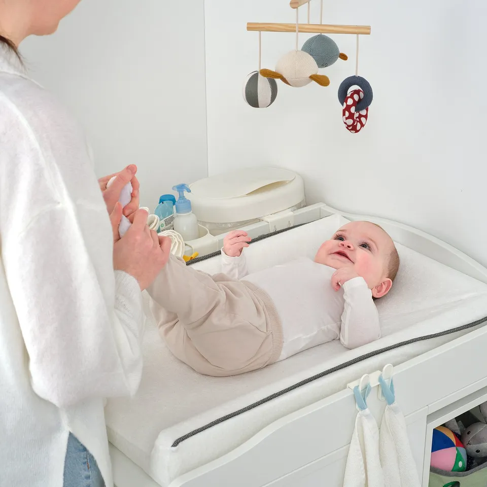 Adult attending baby on changing table with essentials and mobile.