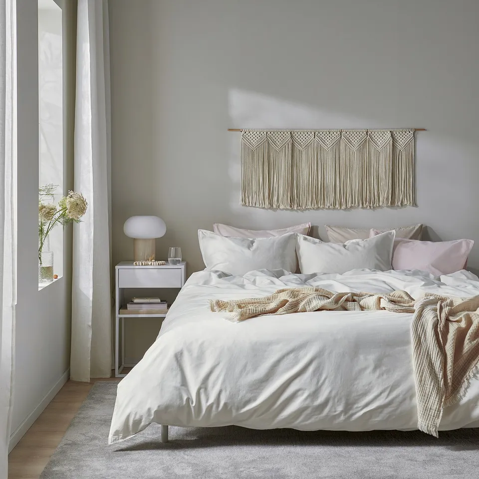 Modern bedroom with ÄNGSLILJA duvet, pillows, macrame wall art, bedside table, lamp, books. Calm, minimalist decor.