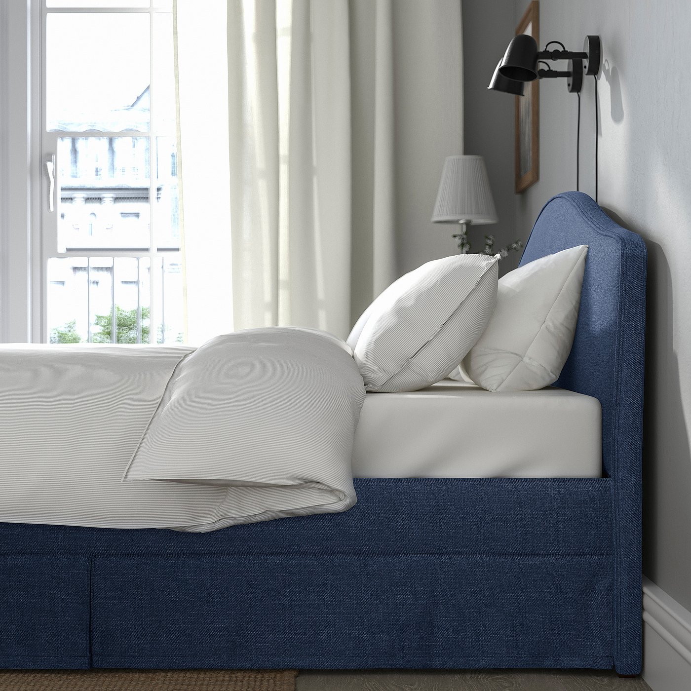 Bed with soft headboard, white pillows, blue cover. Wall light beside, window with sheer curtains.