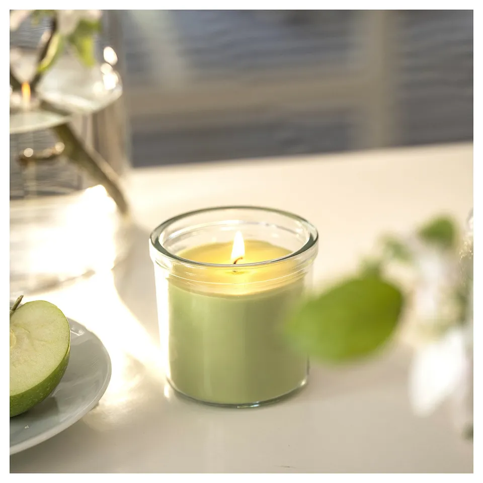 Lit green apple scented candle in glass on table with plate and fruit.