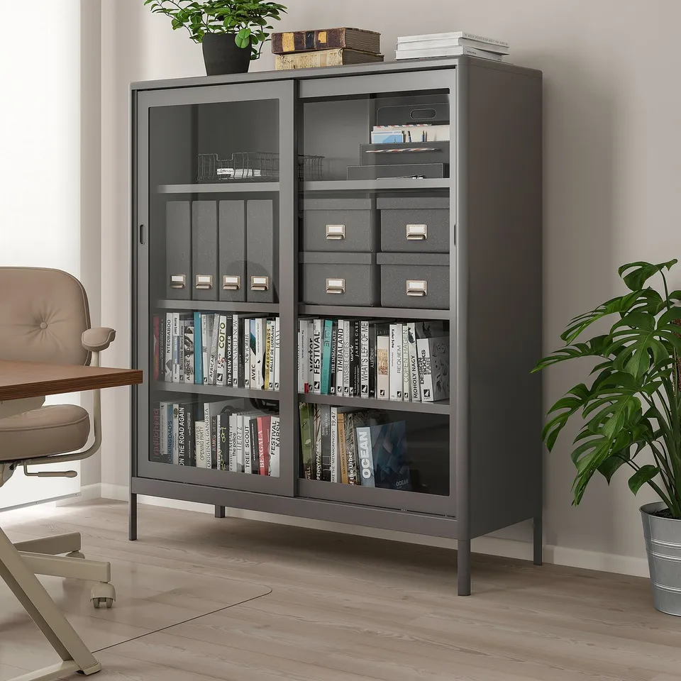 Black IDÅSEN cabinet with glass doors and metal frame, holding books and decor, taller than nearby chair and plant.