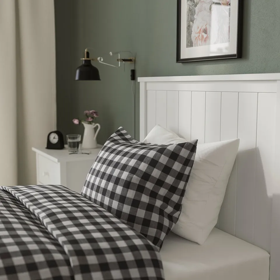 White bed with black-and-white plaid bedding, two pillows, bedside table with lamp, flowers, and framed picture on wall.