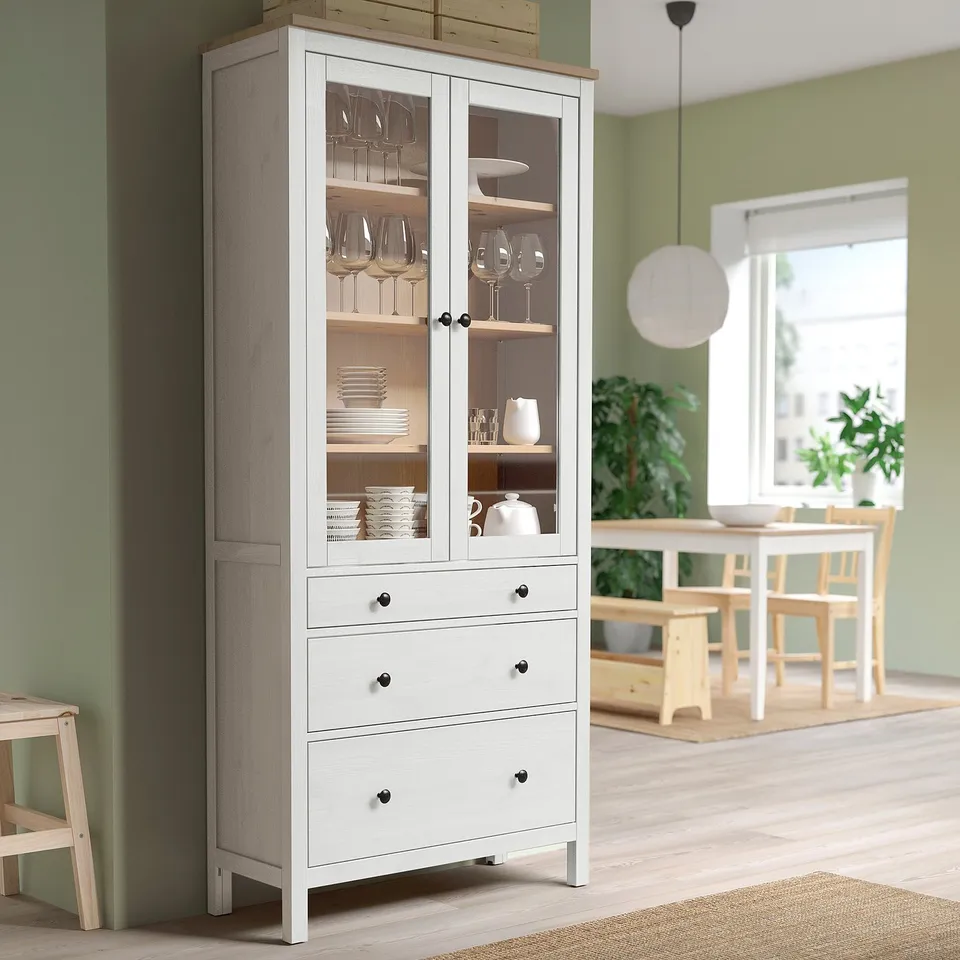 HEMNES wooden cabinet with glass doors and bench against green wall, storing dishes. Background: dining table, chairs, and window.