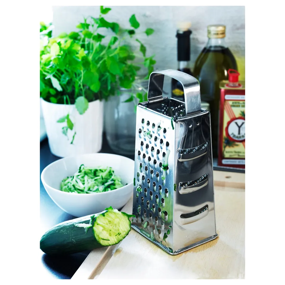Metal cheese grater with a cut cucumber, bowl of grated cucumber, and green herbs on a white countertop.