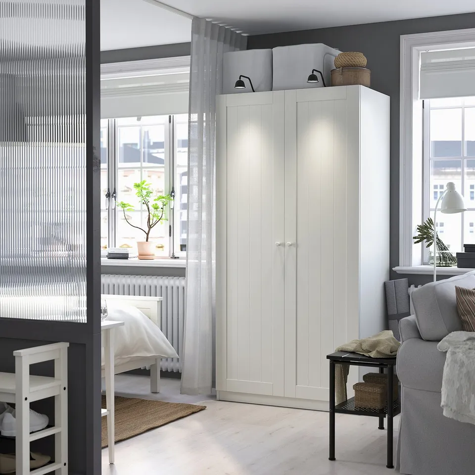Ediated white wardrobe in bright room; near window and bed, taller than chair.
