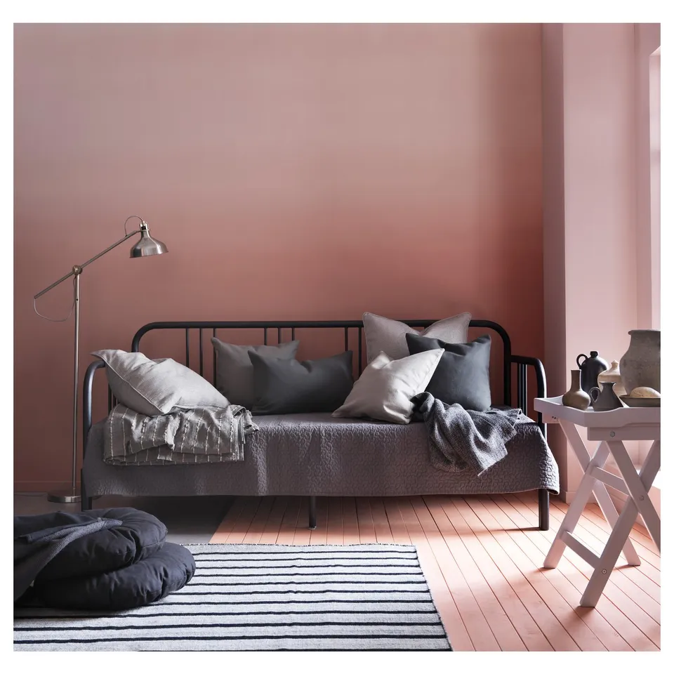 Modern bedroom with FYRESDAL black metal day-bed, grey cushions, pink walls, striped rug, white side table, grey lamp.