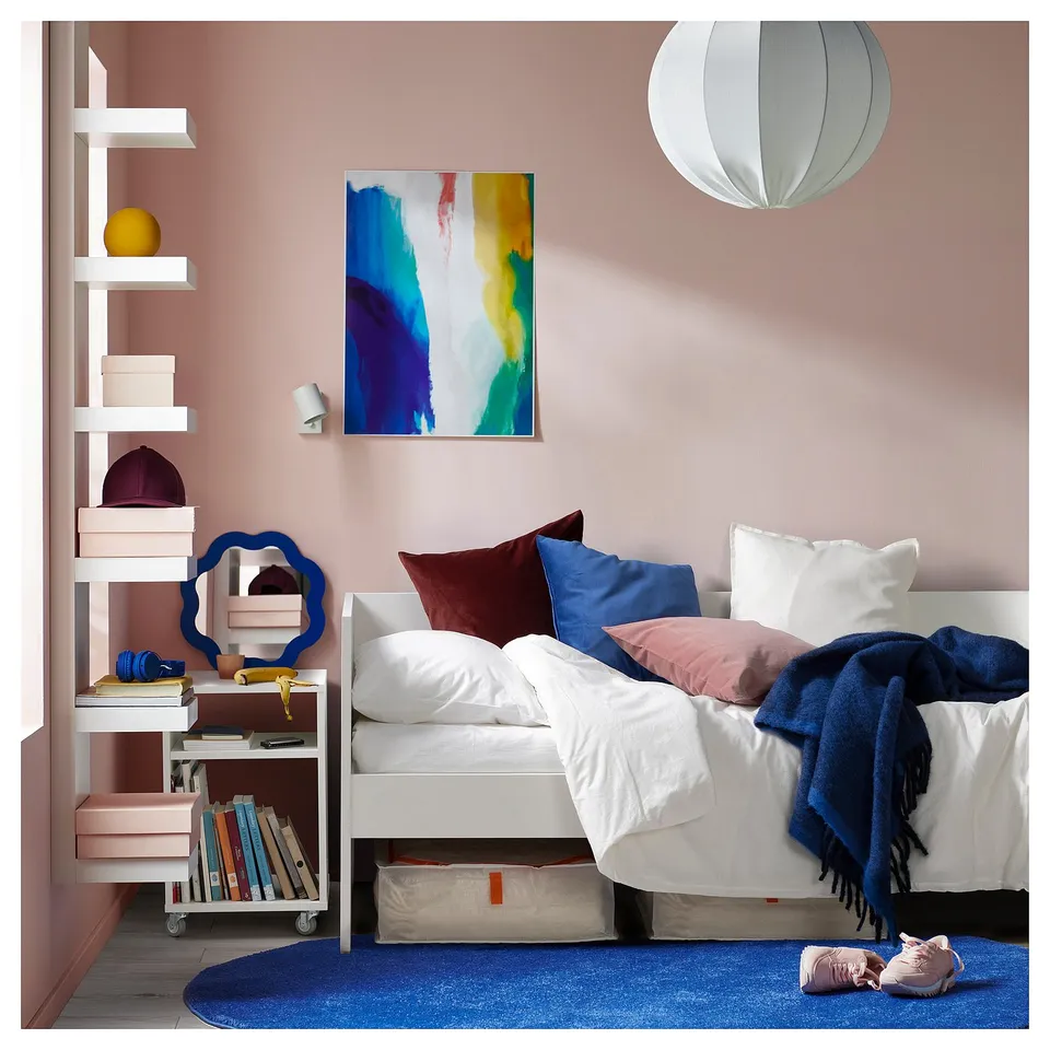 Modern bedroom with pink walls, white daybed, colourful pillows, blue rug, shelves with books, and abstract painting.