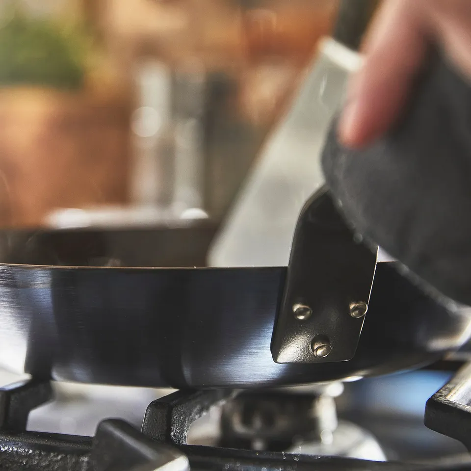 Hand lifts seasoned carbon steel pan by handle, placing it on stovetop burner.