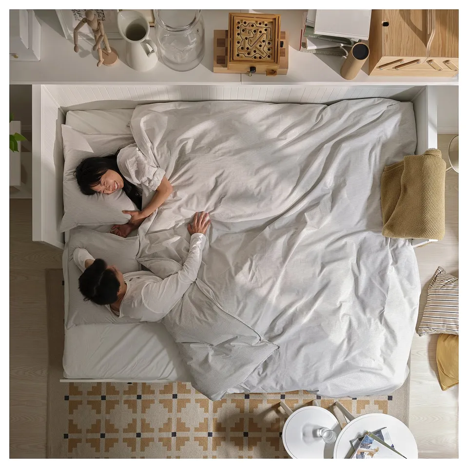 A couple sleeps on a grey HEMNES day-bed. The room has wooden floors, a patterned rug, and decor items on shelves.