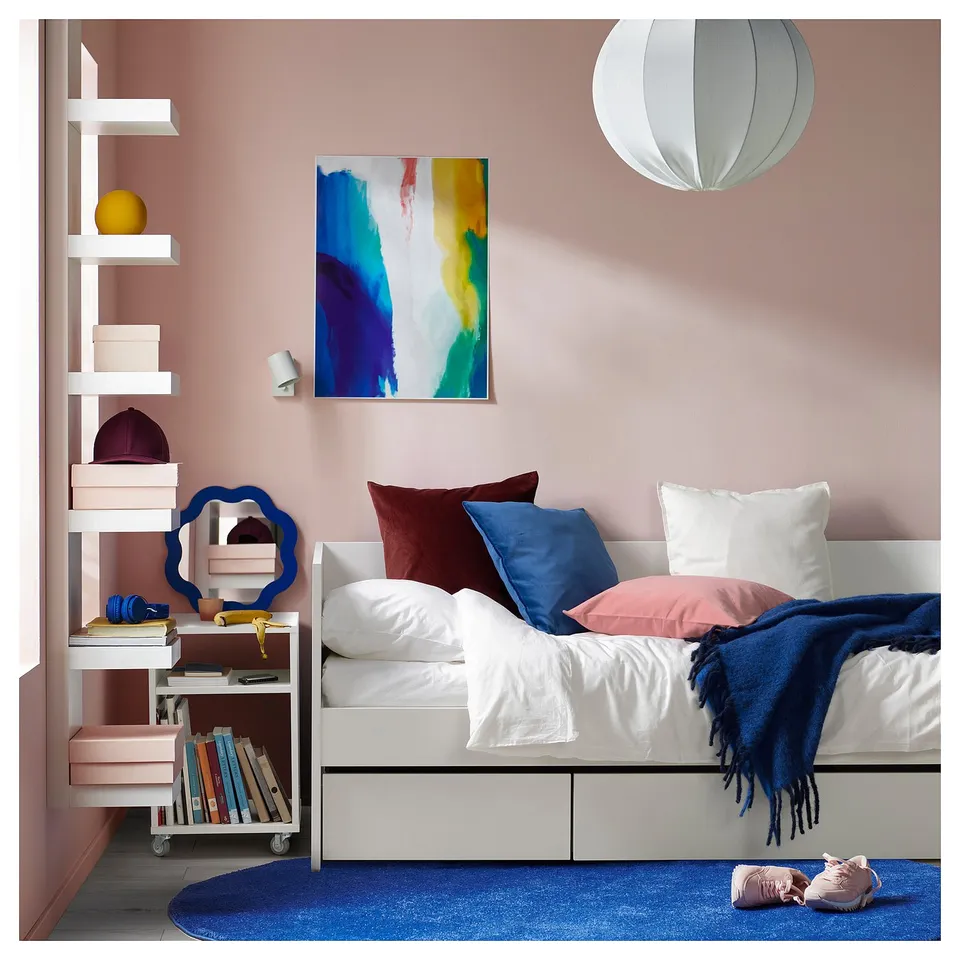 Bedroom with pink walls, white daybed, blue accents, colourful art, floating shelves with books, and a blue rug.