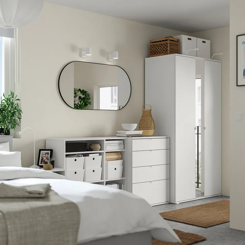 Modern bedroom with white furniture: bed, drawers, wardrobe, and mirror. Natural accents, woven baskets, and plants for decor.