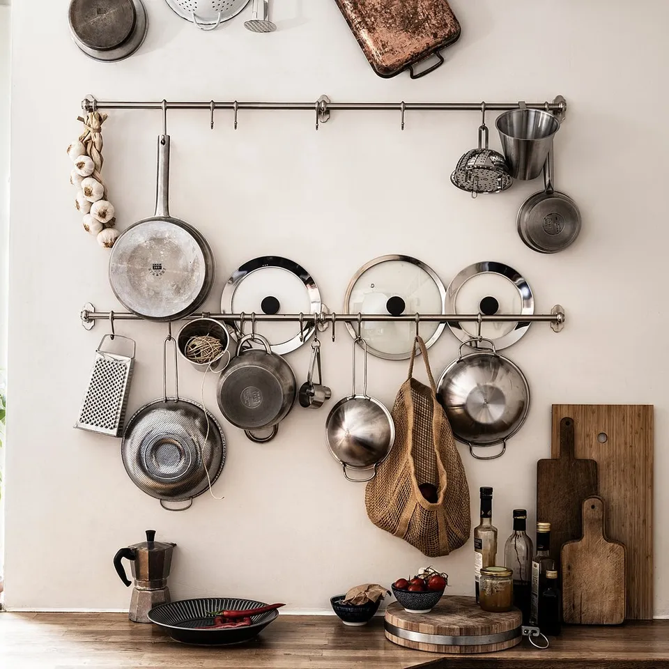 Kitchen wall with hanging pots, pans, and utensils. Stainless steel and wooden items, white wall, green plant.