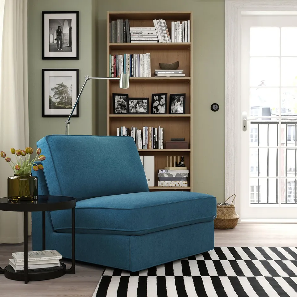 Cosy blue KIVIK sofa-bed in modern living room. Near bookshelf, striped rug, and vase with flowers.