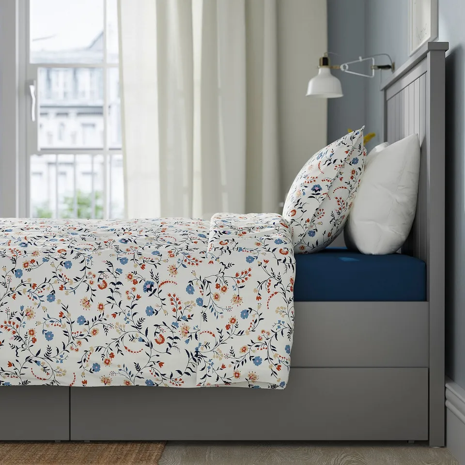 Bed with floral sheets, 2 drawers, wall lamp, city view.