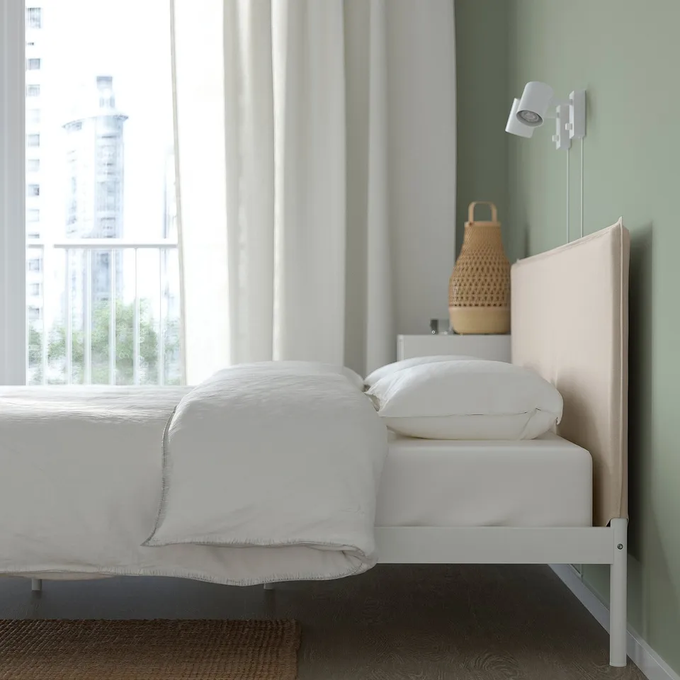Modern white bed frame with upholstered headboard, positioned near a window with city view. Clean design, visible screws, and stable headboard.