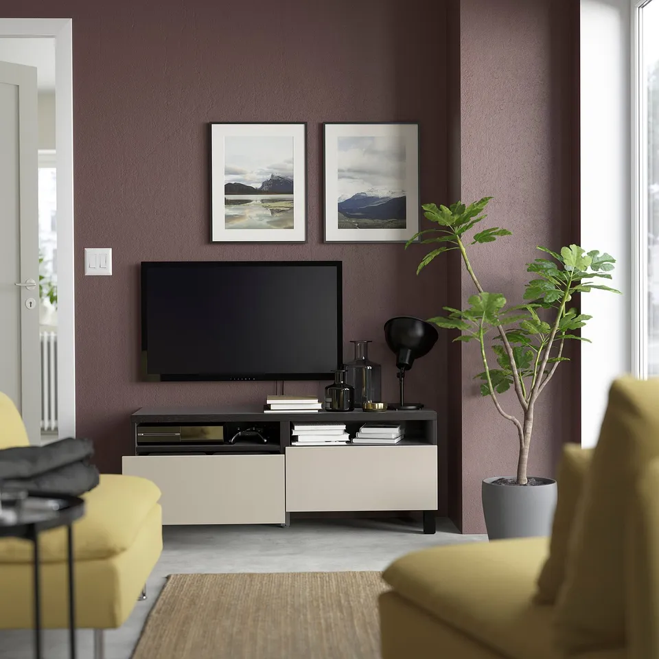 Modern living room with purple walls, featuring BESTÅ tv unit, tv, books, decor, and mountain prints. Yellow sofas flank a rug.