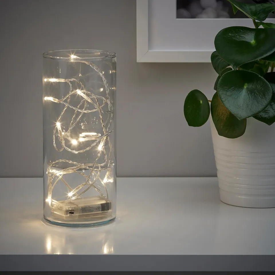 Clear glass cylinder with warm led lights inside, sits on white shelf beside a potted plant.