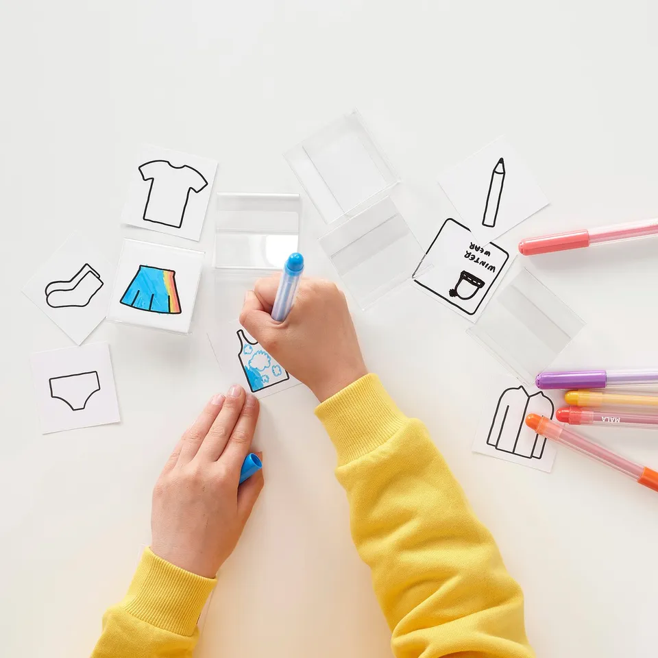 A child in a yellow sweater decorates clothing labels with markers. Art supplies and paper lay on a white surface.