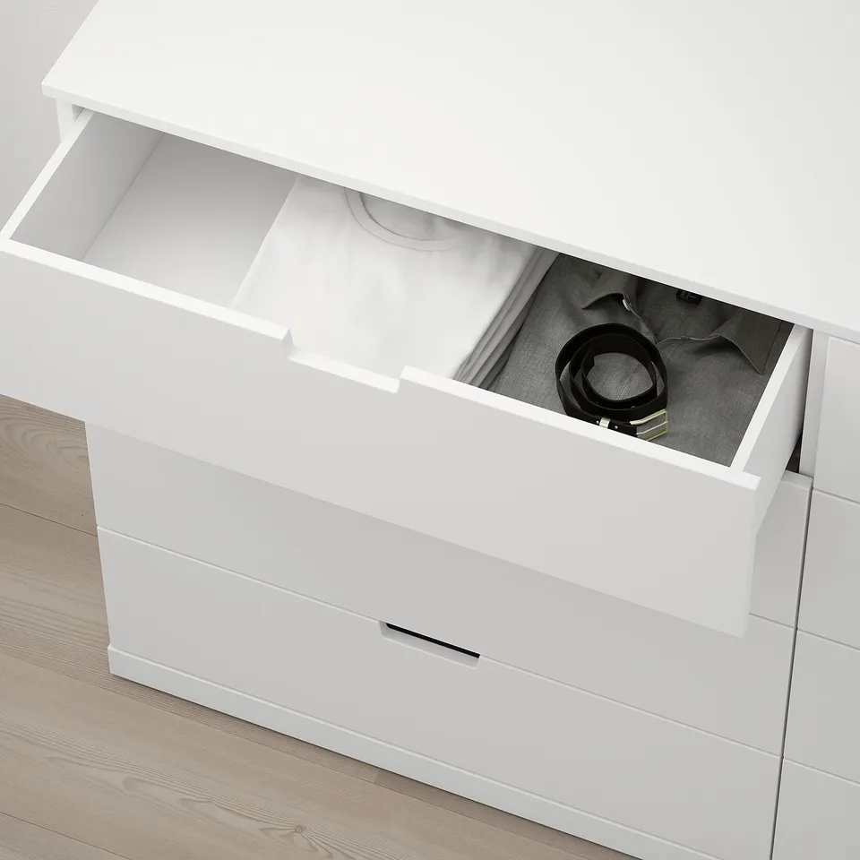 Open white NORDLI dresser with visible drawer contents, including a grey sweater and coiled cables, resting on wooden floor.
