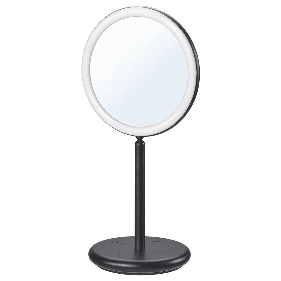 An EKFÄNN two-sided table mirror. Its black, circular with a pinched middle, and integrated lighting. The design includes a stand with a base.