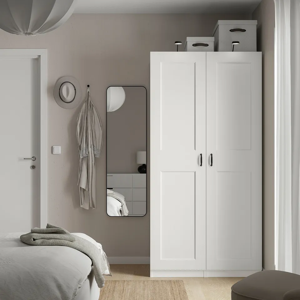 Minimalist bedroom with white PAX wardrobe, mirror, hanging hat, and partially visible bed. Wardrobe is tall and compact, fitting neatly beside door.