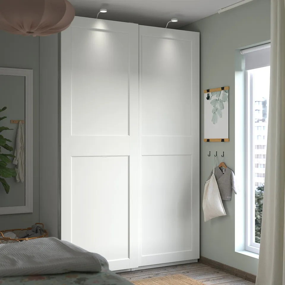 Large white wardrobe with sliding doors in bedroom corner, next to window with curtains, featuring wooden hangers and grey clothes.