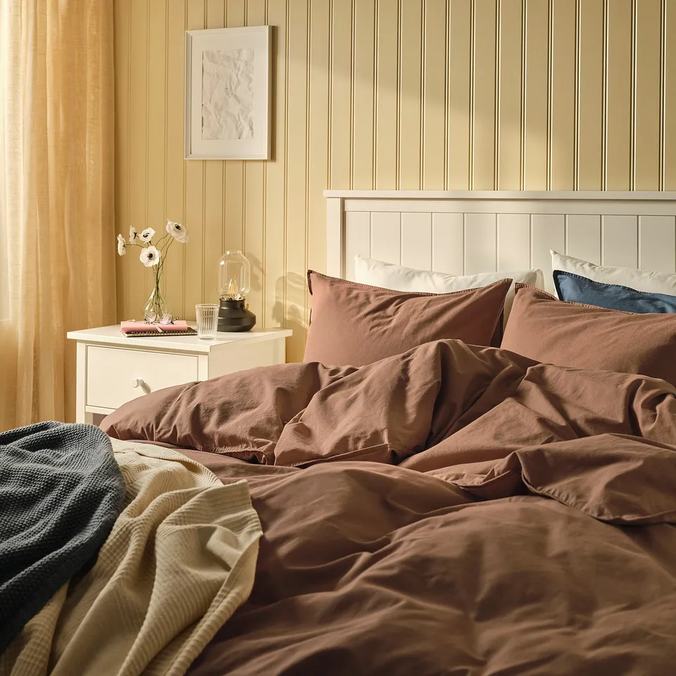 Bed with ÄNGSLILJA duvet, recycled cotton, pillows, headboard, bedside table with flowers and lamp.