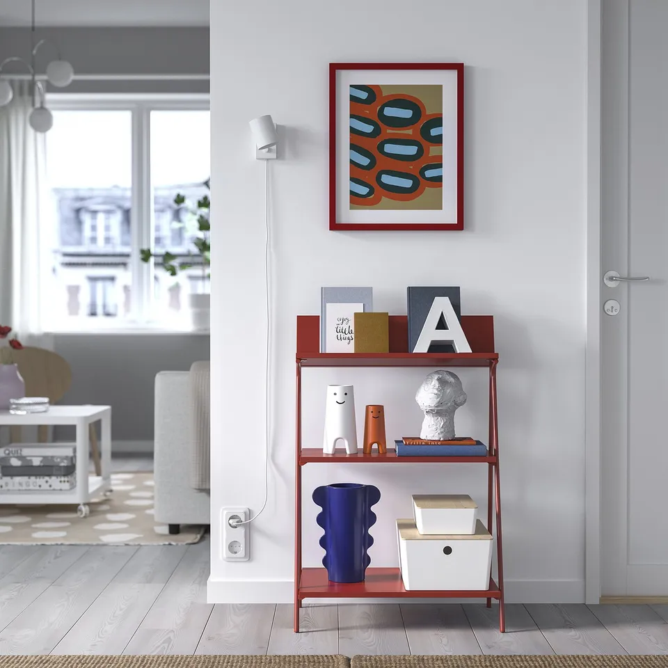 Red KRÖNIKÖR shelf displays books, vase, toys, near white door. Bright painting above.