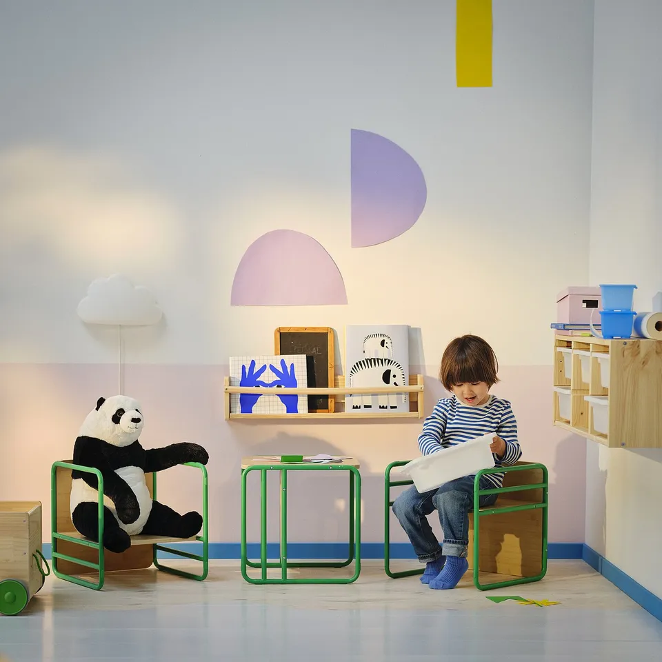 Kids playroom with SMÅSKRAKE chair-table, panda toy, books, and wall decals. Child sits drawing on green, bamboo chair.