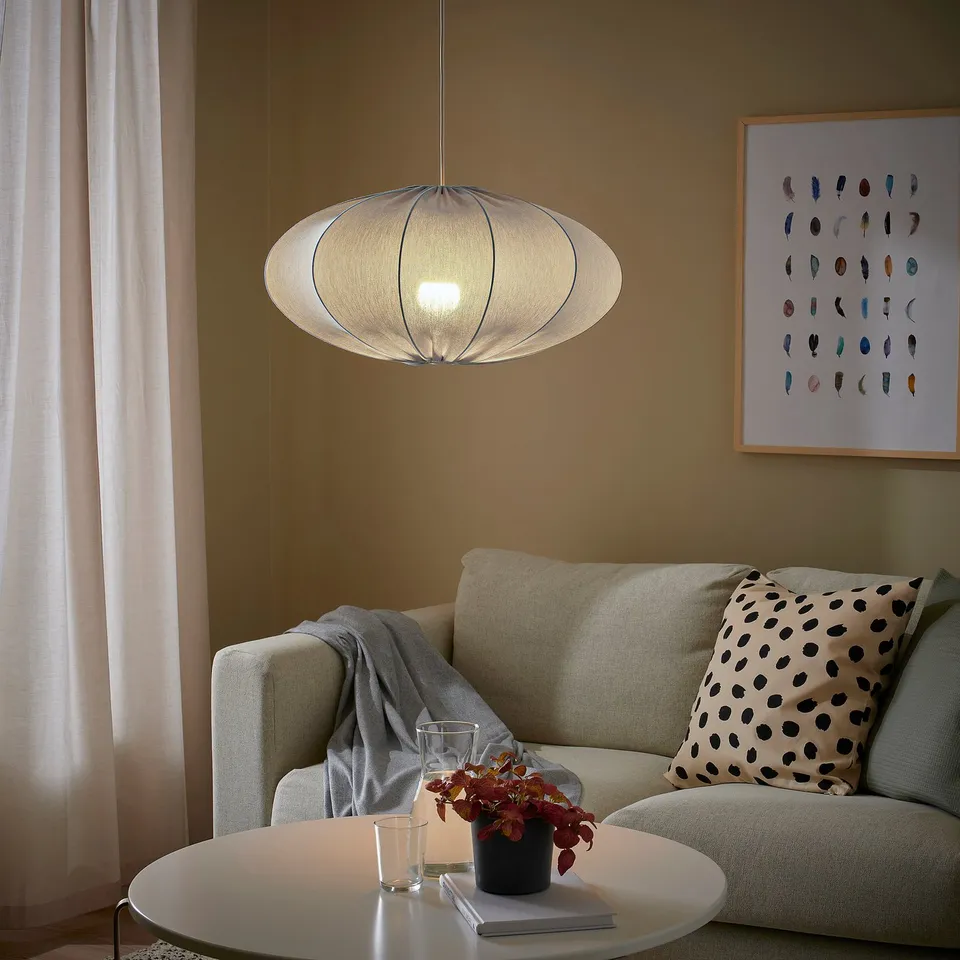 Modern living room with REGNSKUR lamp, beige sofa, pillows, blanket, coffee table, book, plant, and colourful feather art.