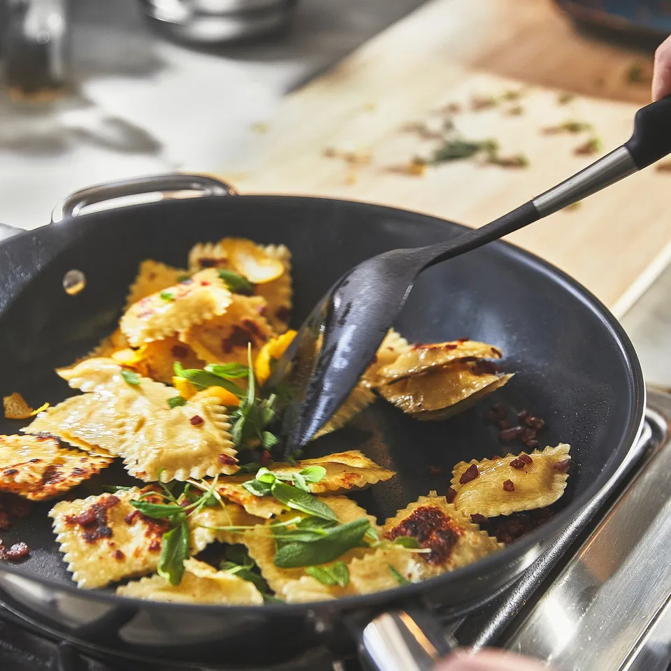 A black frying pan on a stove cooks ravioli, topped with green herbs. Two handles and rounded interior are visible.