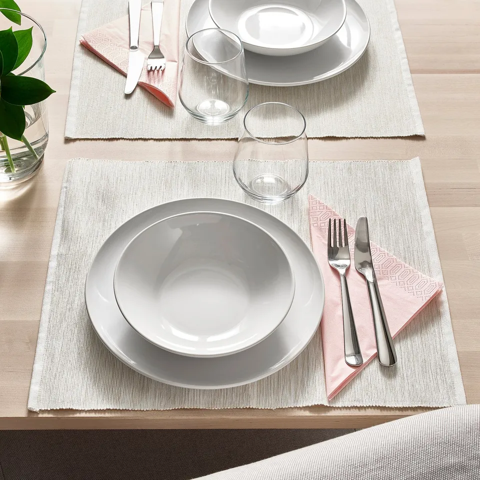Two place settings with white plates, glasses, forks, knives, and pink napkins on woven placemats on a wooden table.